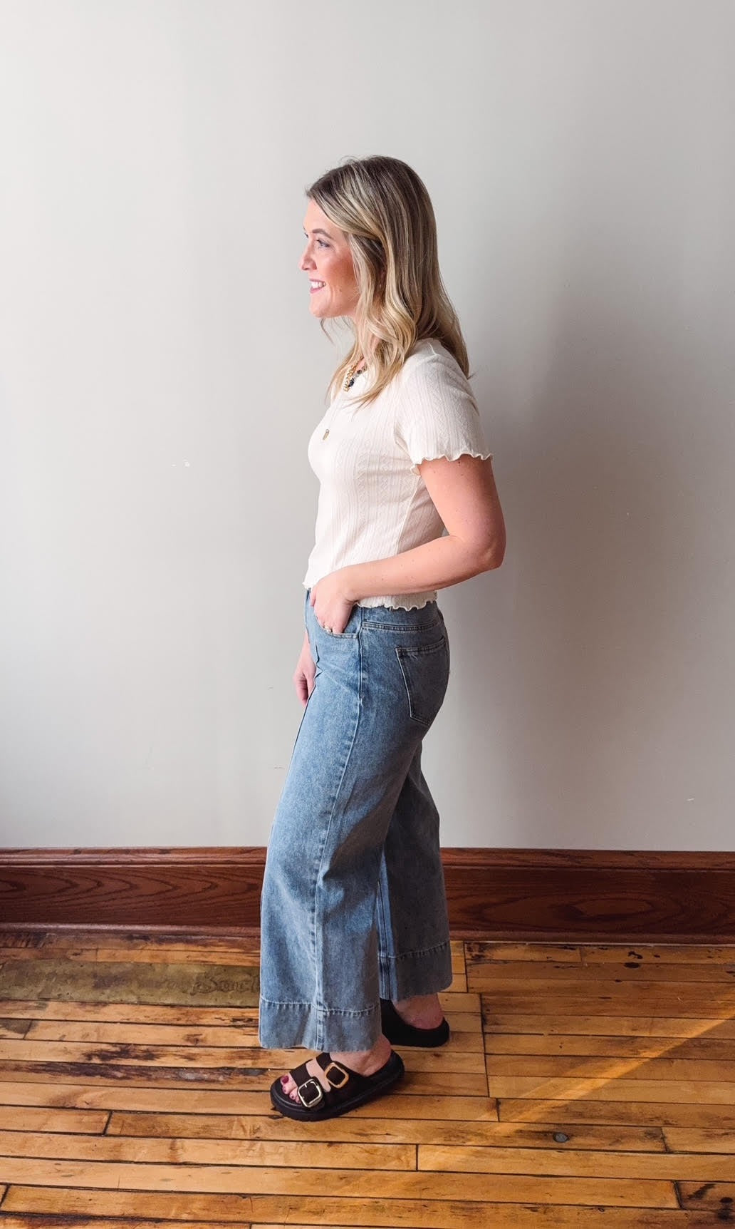 Woman wearing a white shirt and blue jeans standing against a plain wall.