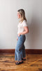 Woman wearing a white shirt and blue jeans standing against a plain wall.