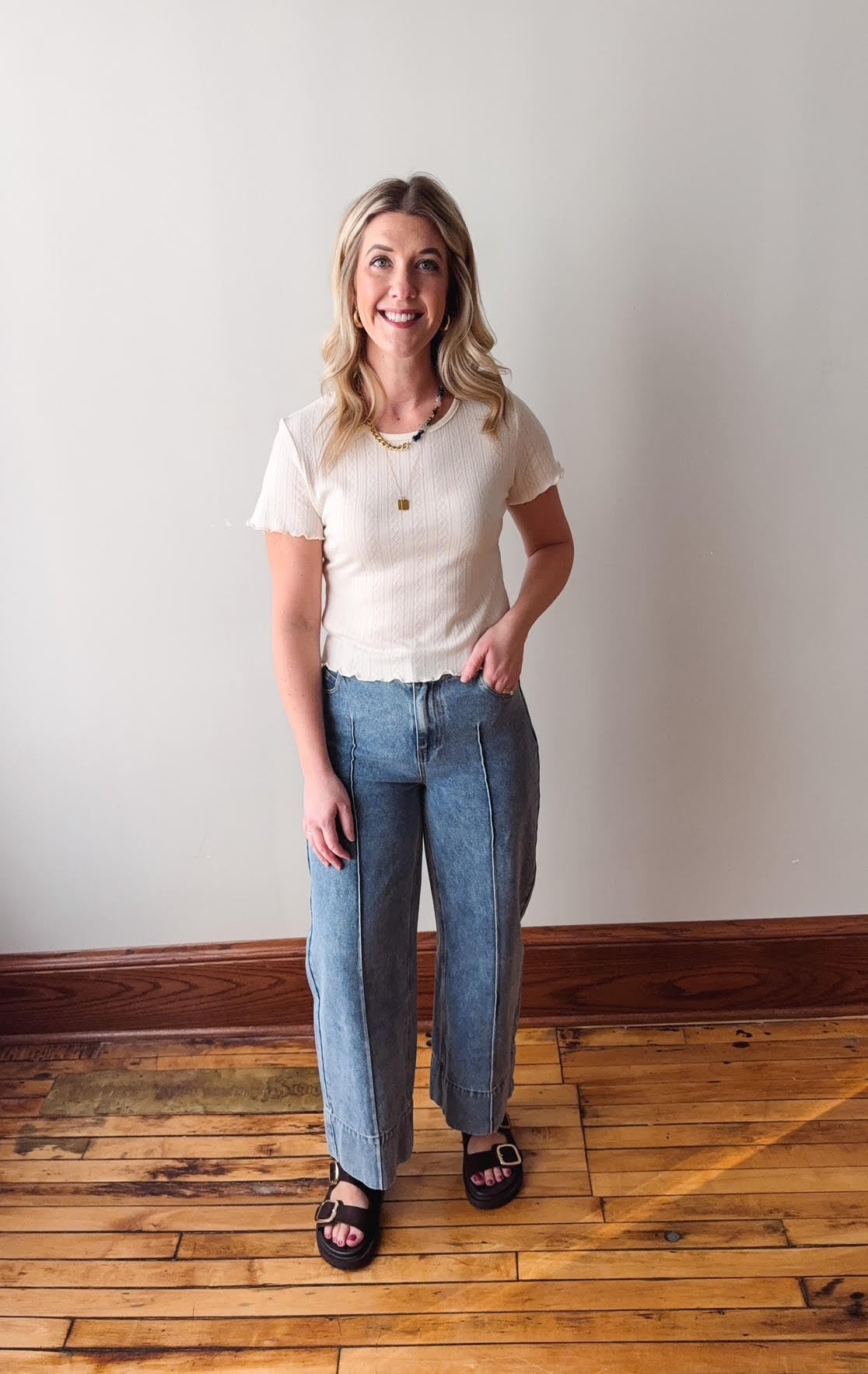 Woman wearing a white t-shirt and blue jeans standing on a wooden floor with a plain wall background