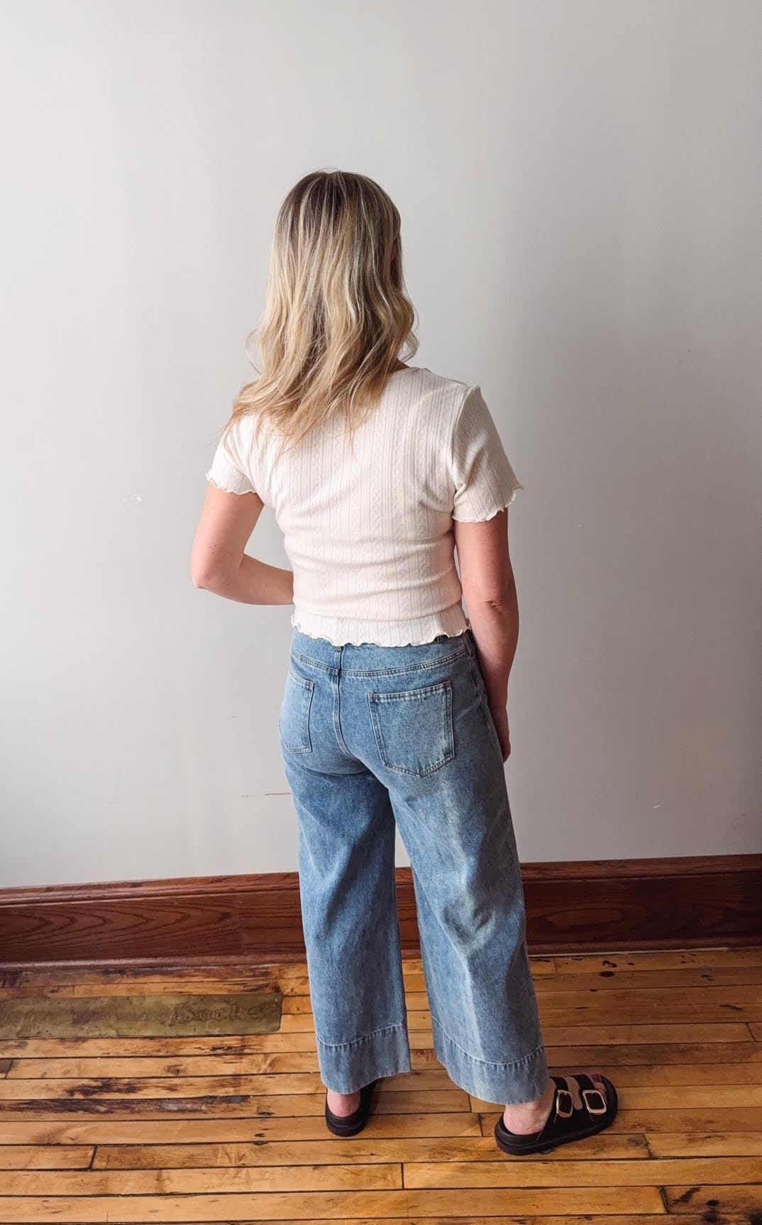 Person wearing a white top and blue jeans standing on a wooden floor with a plain wall background