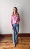 Woman wearing a pink cardigan and blue jeans standing on a wooden floor against a white wall.