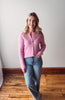Woman wearing a pink sweater and blue jeans standing in a room with wooden flooring and white walls.