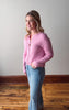 Woman wearing a pink sweater and blue jeans standing in a room with wooden flooring and a white wall.