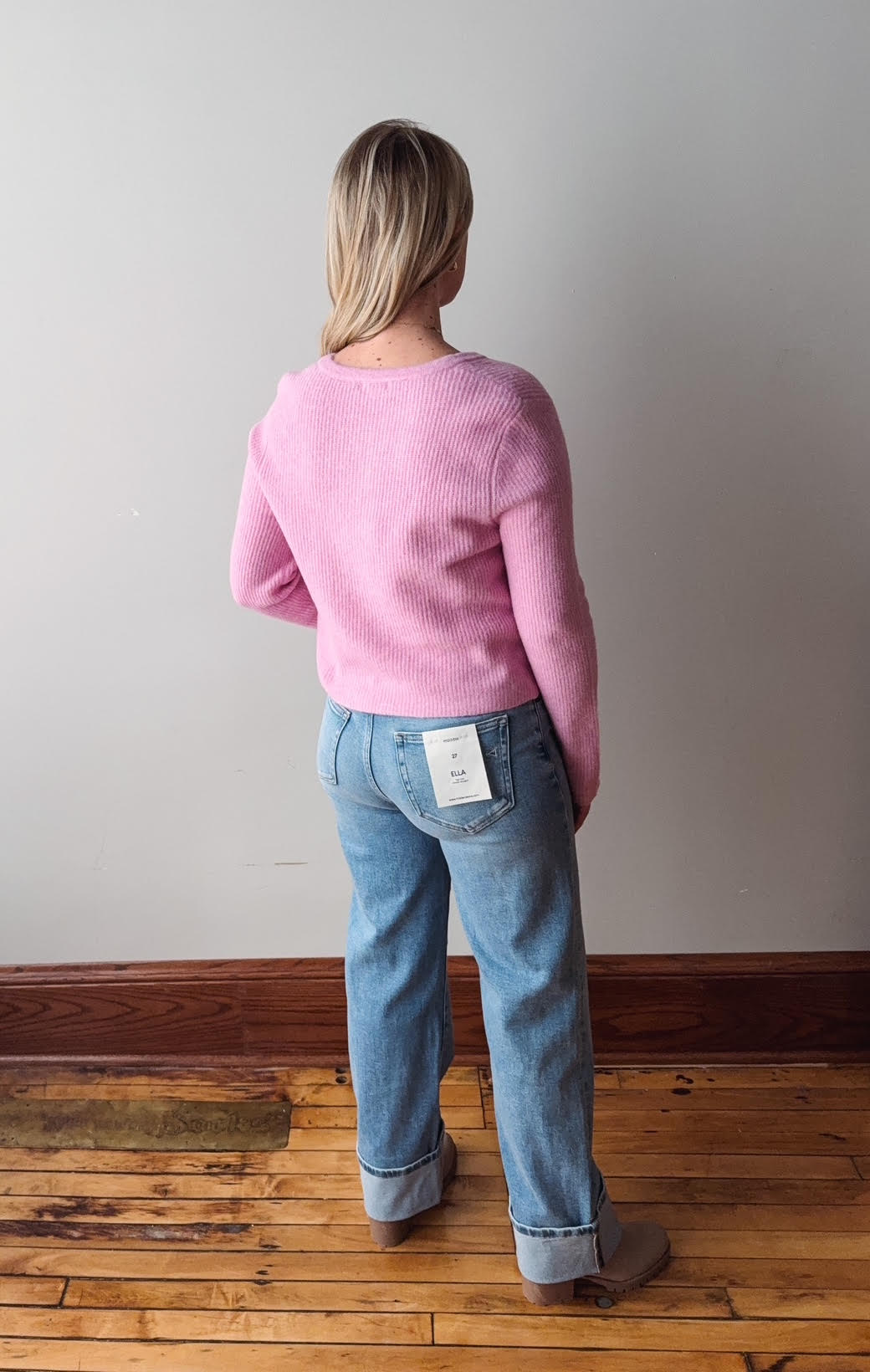 Person wearing a pink sweater and blue jeans standing on a wooden floor with a plain wall background