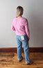 Person wearing a pink sweater and blue jeans standing on a wooden floor with a plain wall background