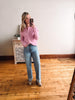 Woman taking a mirror selfie wearing a pink shirt and blue jeans in a room with wooden flooring.