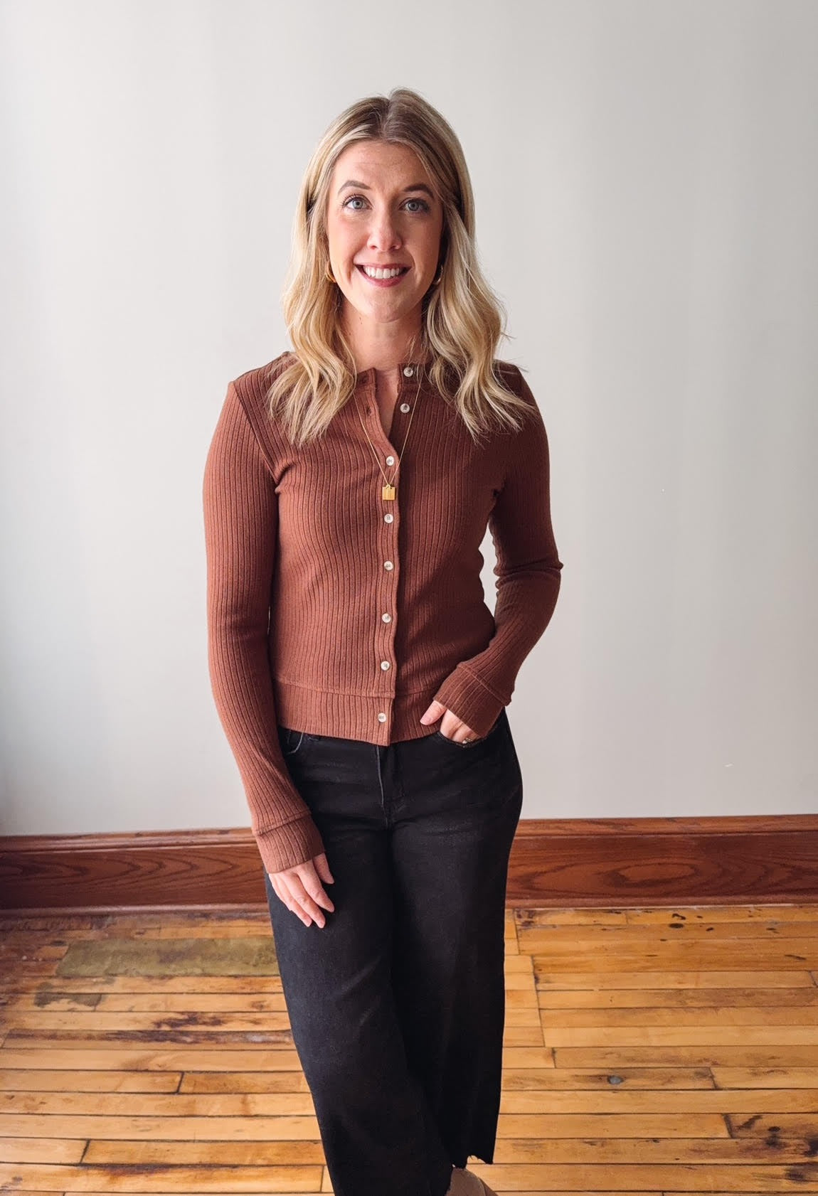 Woman wearing a brown button-up shirt and black pants standing in a room with wooden flooring and a white wall.