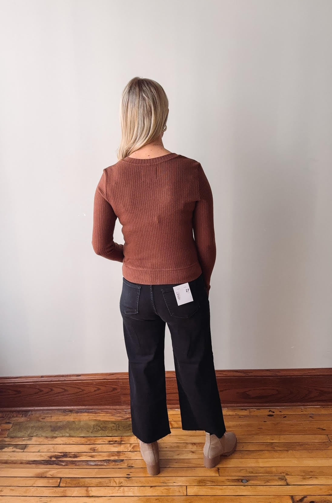 Person wearing a brown sweater and black pants standing on a wooden floor with a white wall background