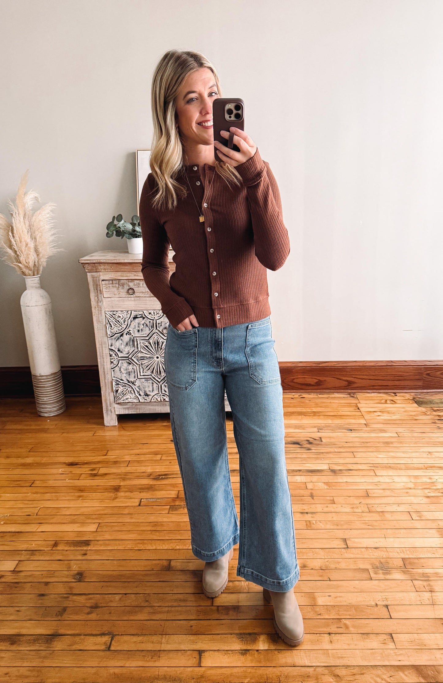 Person wearing a brown shirt and blue jeans taking a mirror selfie in a room with wooden flooring and a decorative cabinet.