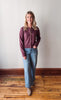 Woman wearing a maroon shirt and blue jeans standing on a wooden floor with a white wall background