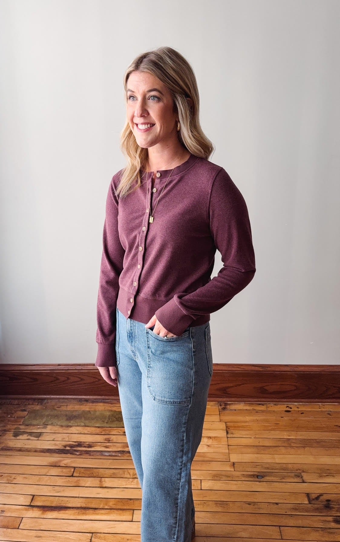 Woman wearing a maroon long-sleeve shirt and light blue jeans standing on a wooden floor.