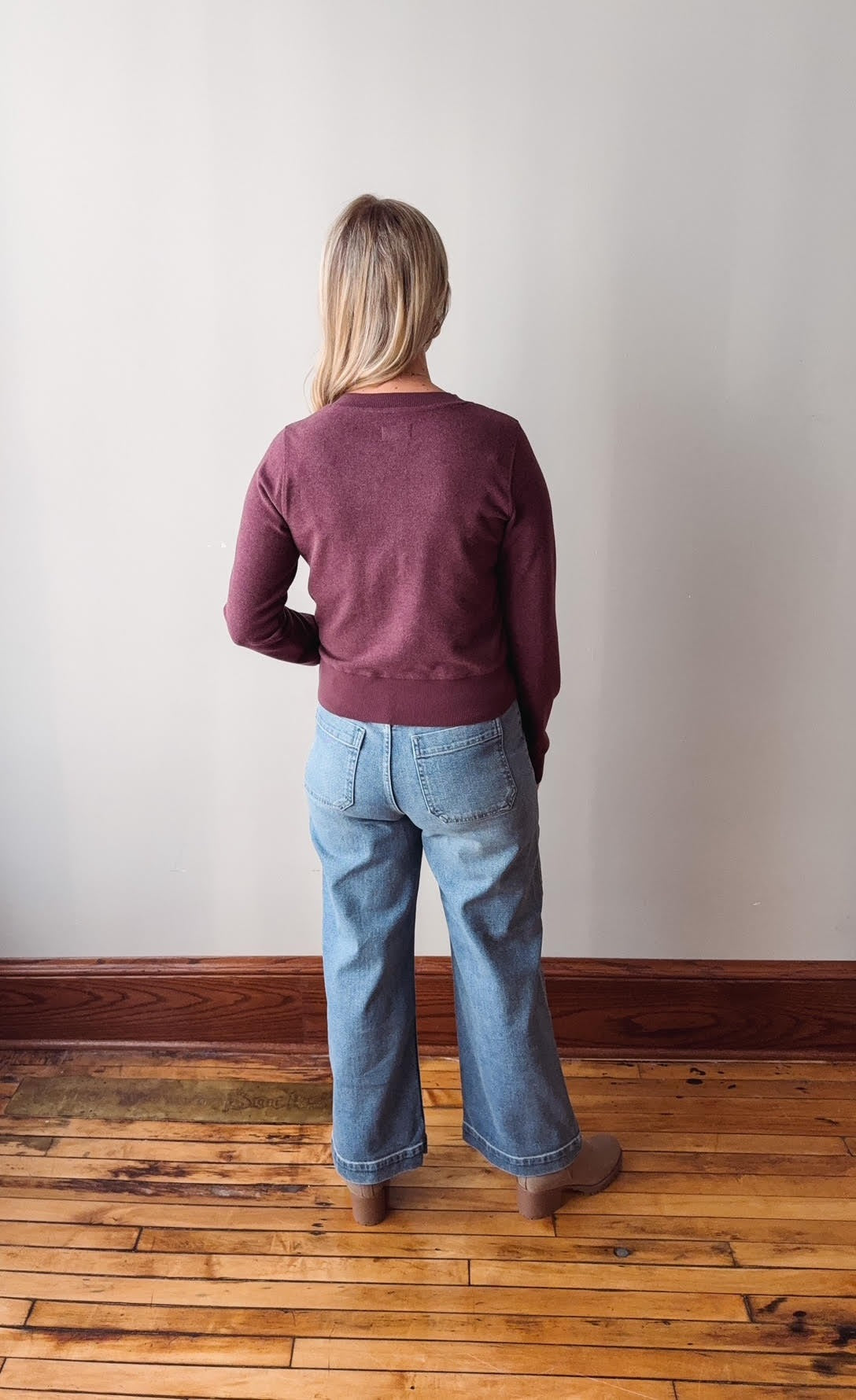 Person wearing a maroon sweater and blue jeans standing in a room with wooden flooring and a white wall.