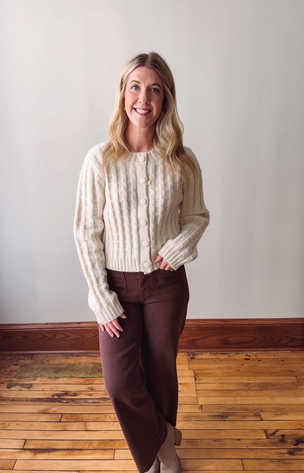 Woman wearing a cream sweater and brown pants standing on a wooden floor with a white wall background