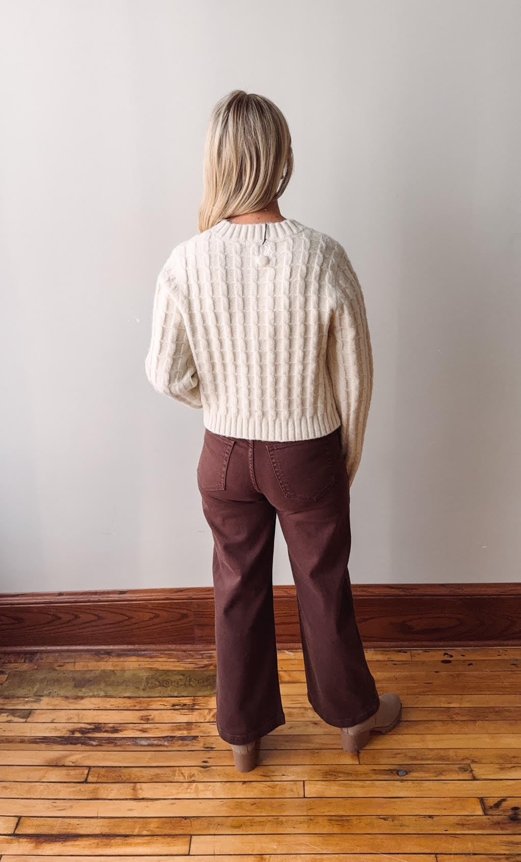 Person wearing a cream sweater and brown pants standing on a wooden floor against a white wall.