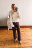 Woman taking a mirror selfie wearing a cream sweater and brown pants in a room with wooden flooring.