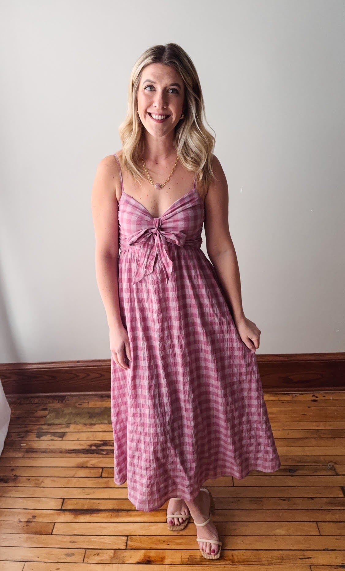 Woman wearing a pink checkered dress standing on a wooden floor with a white wall background