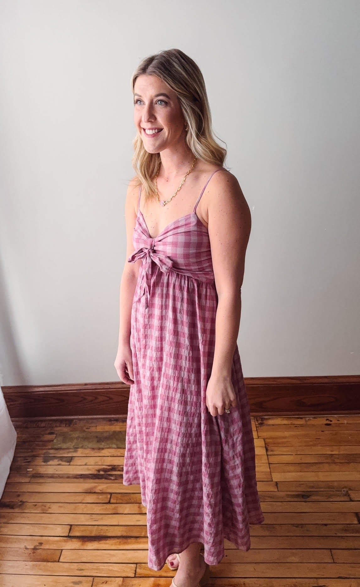 Woman wearing a pink checkered dress standing on a wooden floor with a white wall background