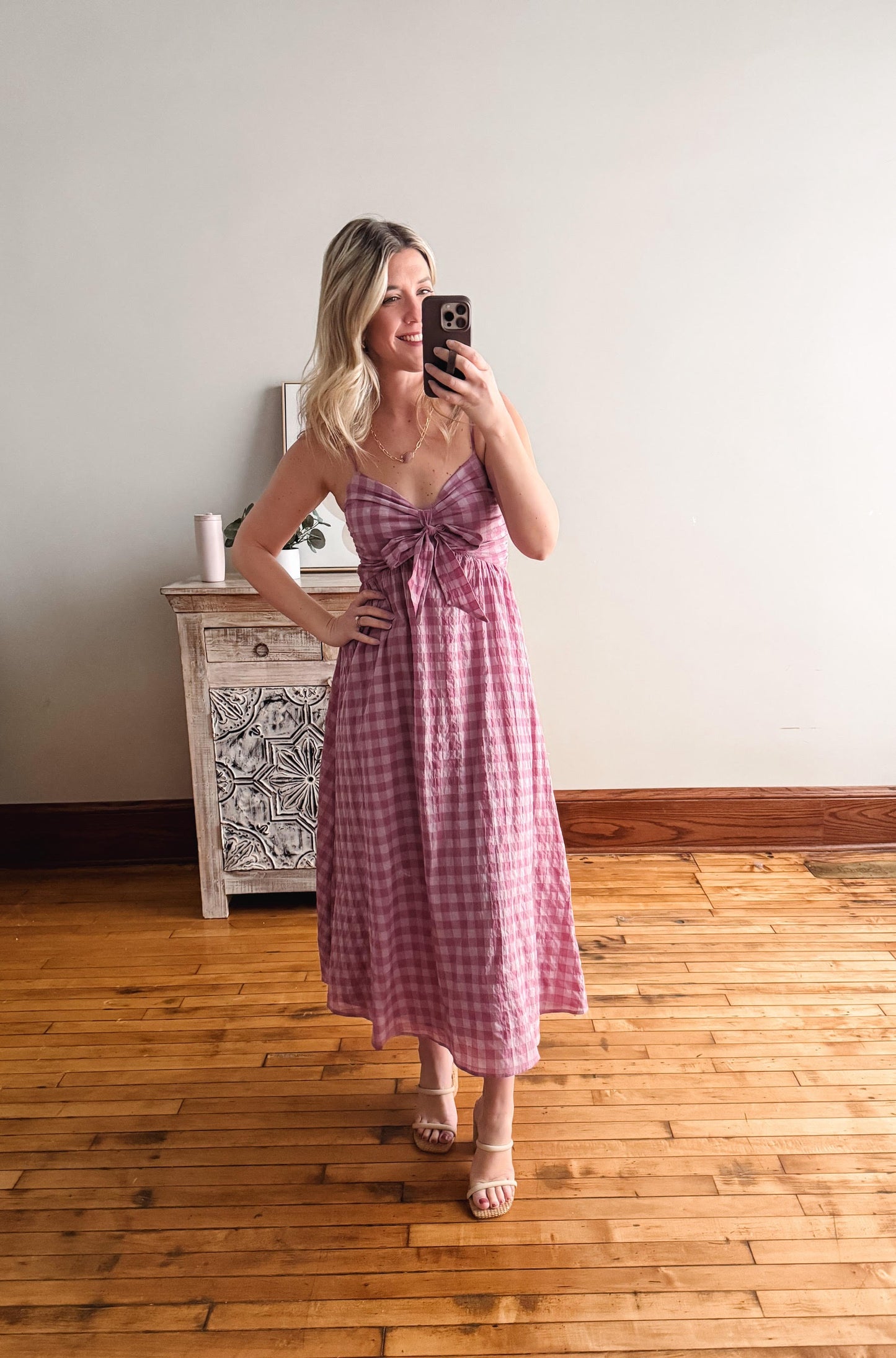 Woman taking a mirror selfie wearing a pink checkered dress in a room with wooden flooring and a white dresser.