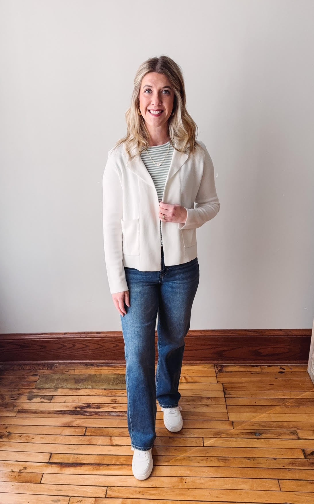 Woman wearing a white jacket, striped shirt, and blue jeans standing on a wooden floor.