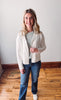 Woman wearing a white blazer, striped shirt, and jeans standing on a wooden floor.
