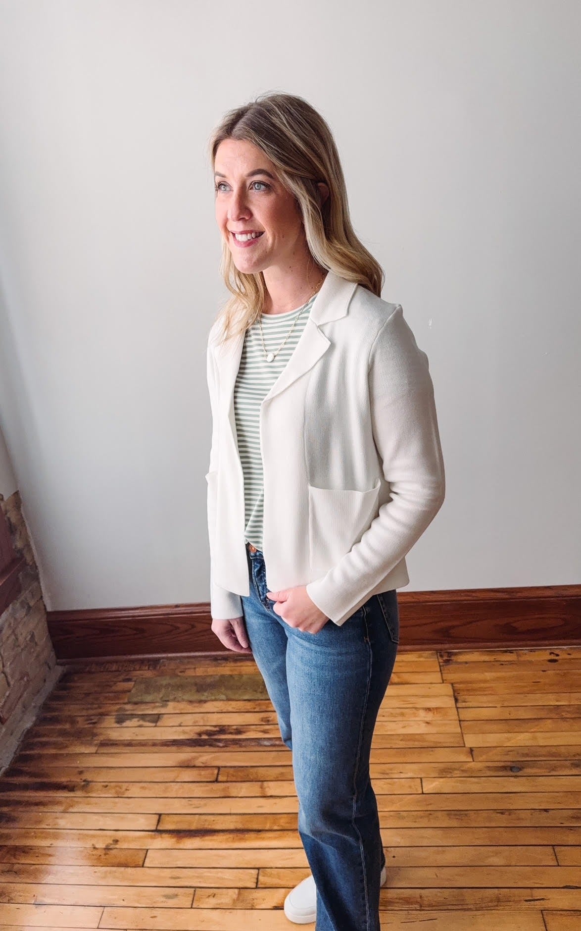 Woman wearing a white blazer over a striped shirt with blue jeans, standing in a room with wooden flooring and a white wall.