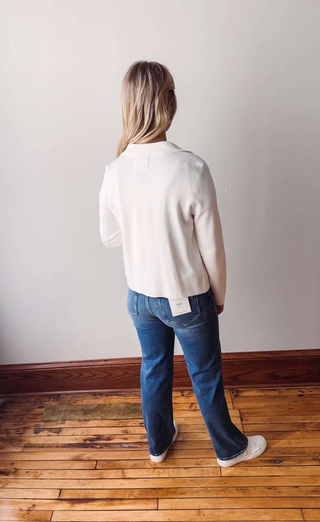 Person wearing a white sweater and blue jeans standing on a wooden floor against a plain wall.