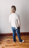 Person wearing a white sweater and blue jeans standing on a wooden floor against a plain wall.