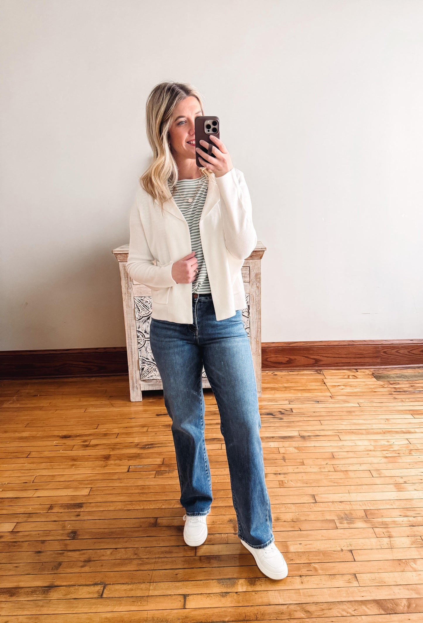 Woman taking a mirror selfie wearing a white cardigan, striped shirt, blue jeans, and white sneakers in a room with wooden flooring.