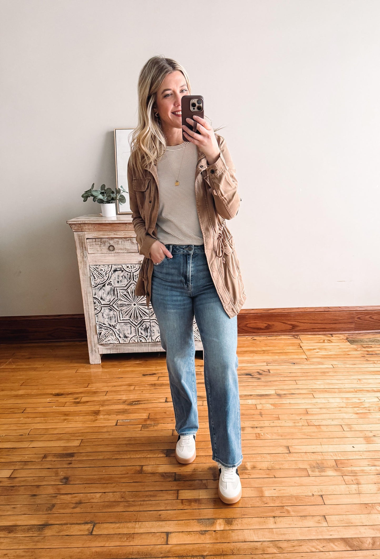 Woman taking a mirror selfie wearing a beige cardigan, white shirt, blue jeans, and white sneakers in a room with wooden flooring.