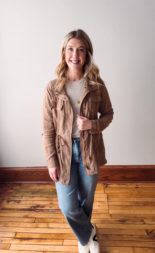 Woman wearing a beige jacket, white shirt, blue jeans, and white sneakers in a room with wooden flooring.