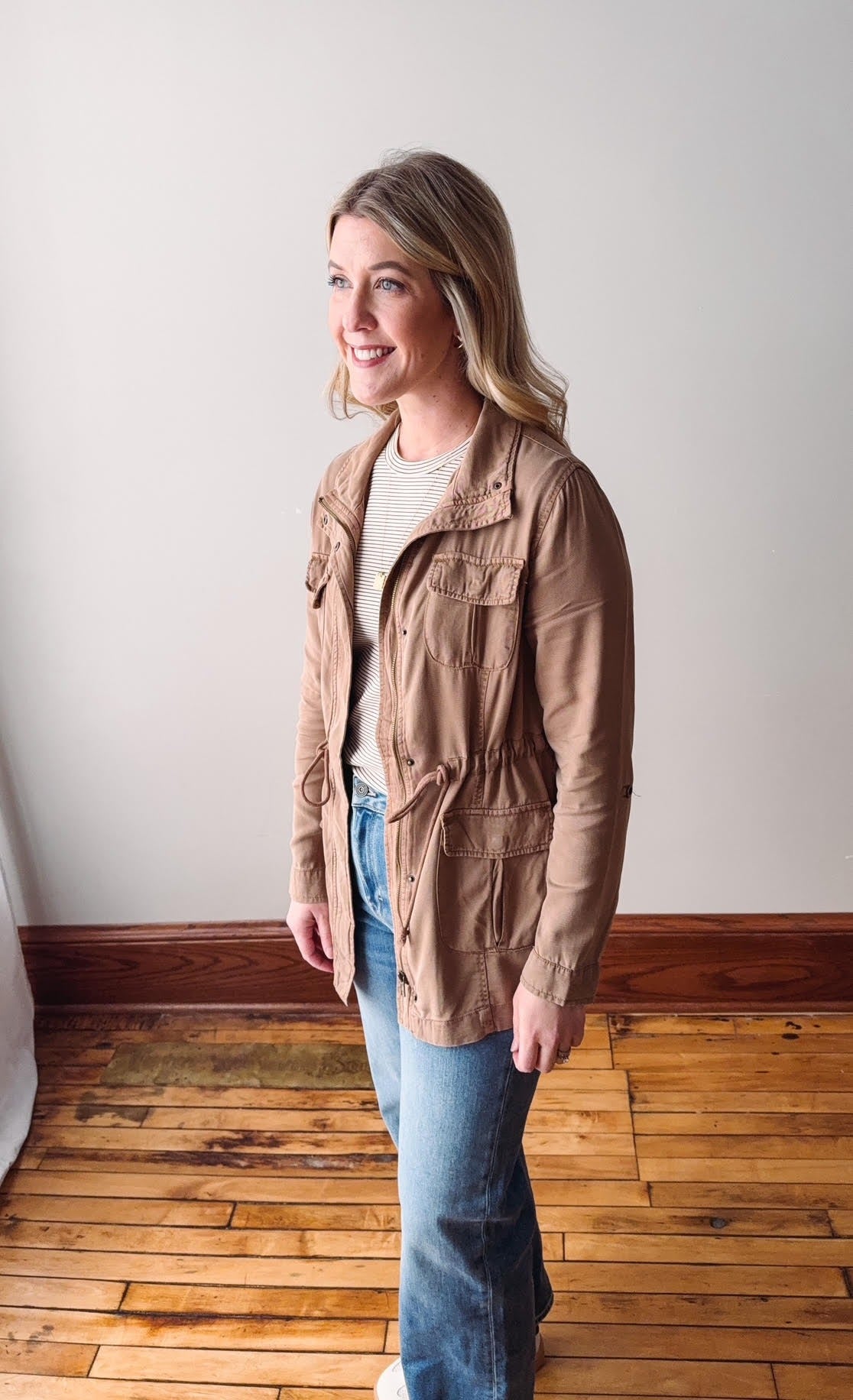 Woman wearing a beige jacket and blue jeans standing on a wooden floor.
