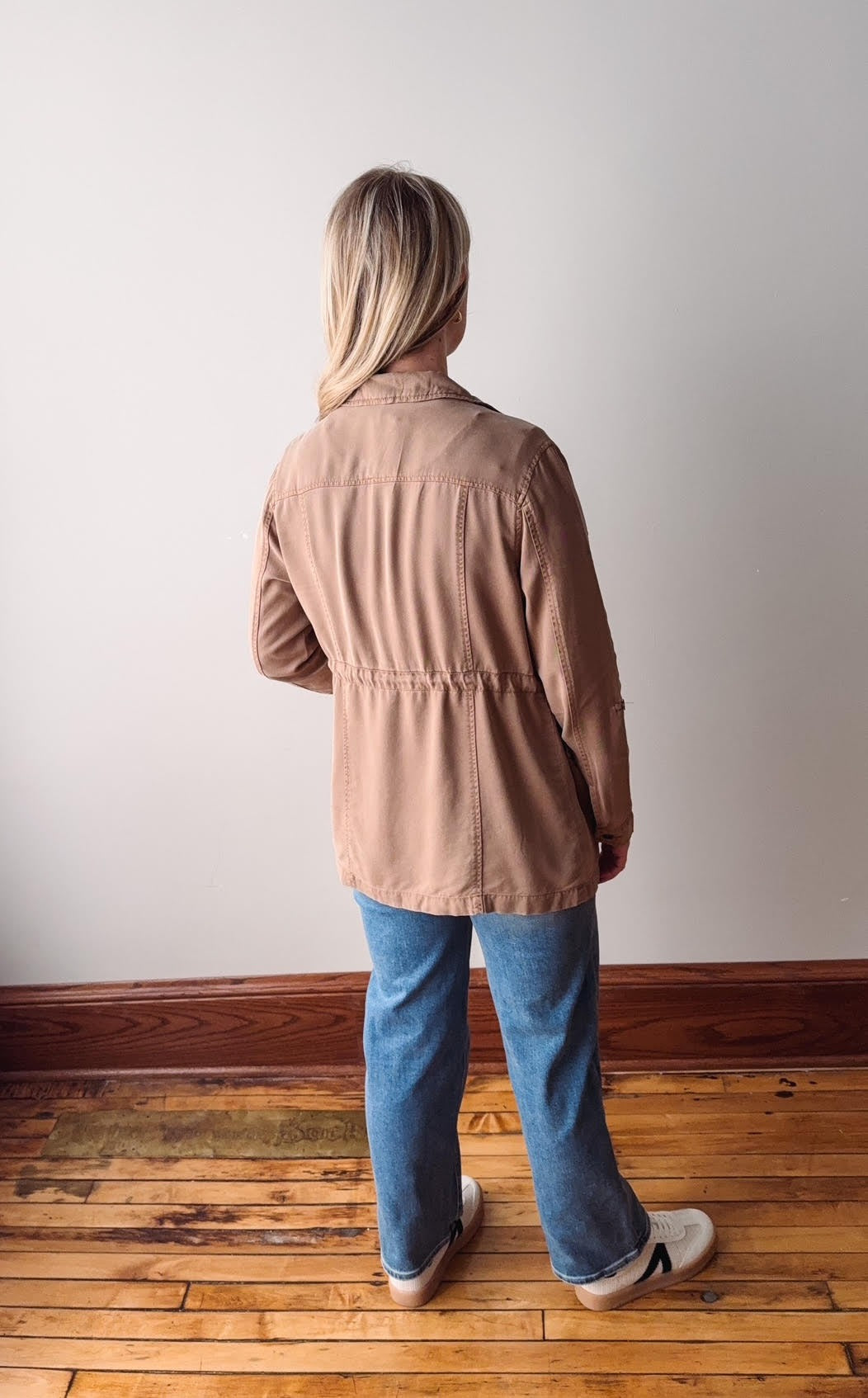 Person wearing a brown jacket and blue jeans standing on a wooden floor against a white wall.
