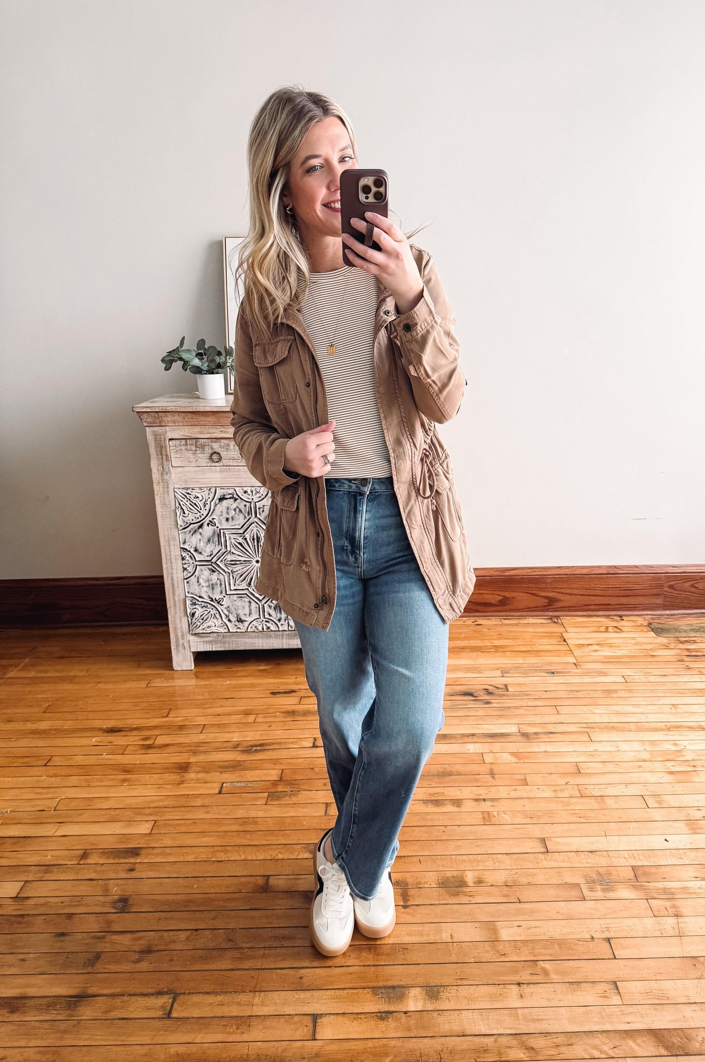 Woman taking a mirror selfie wearing a beige jacket, white shirt, blue jeans, and white sneakers in a room with wooden flooring.