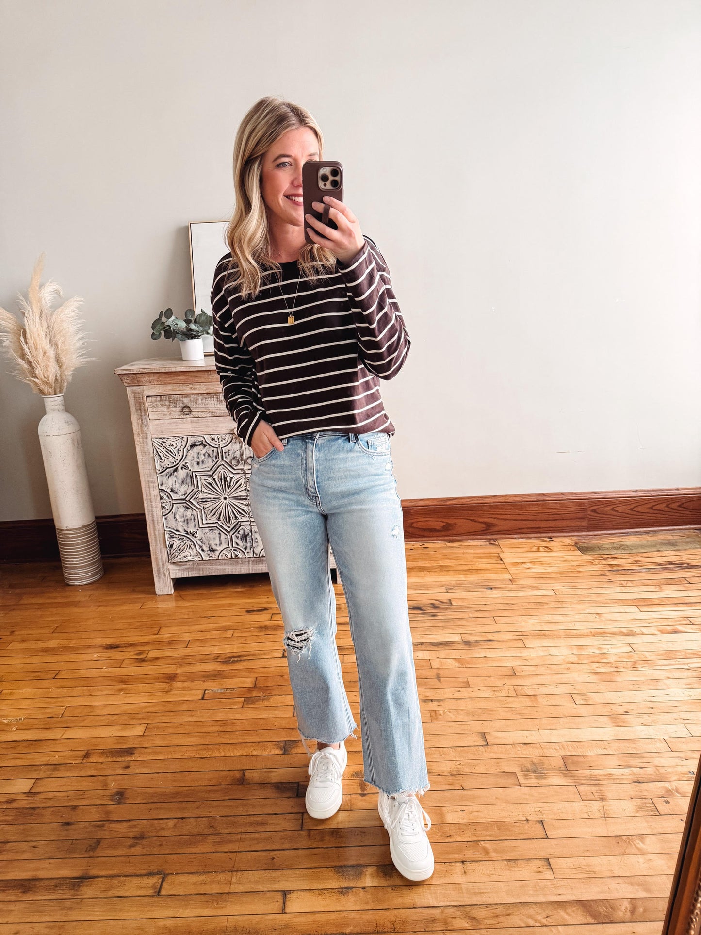 Woman taking a mirror selfie wearing a striped sweater and jeans in a room with wooden flooring.