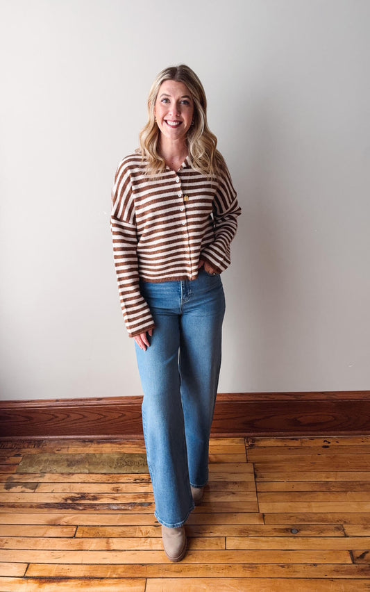Woman wearing a striped sweater and blue jeans standing on a wooden floor against a white wall.