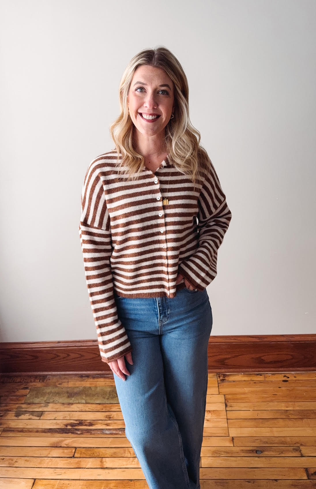 Woman wearing a striped sweater and jeans standing in a room with wooden flooring and a white wall.