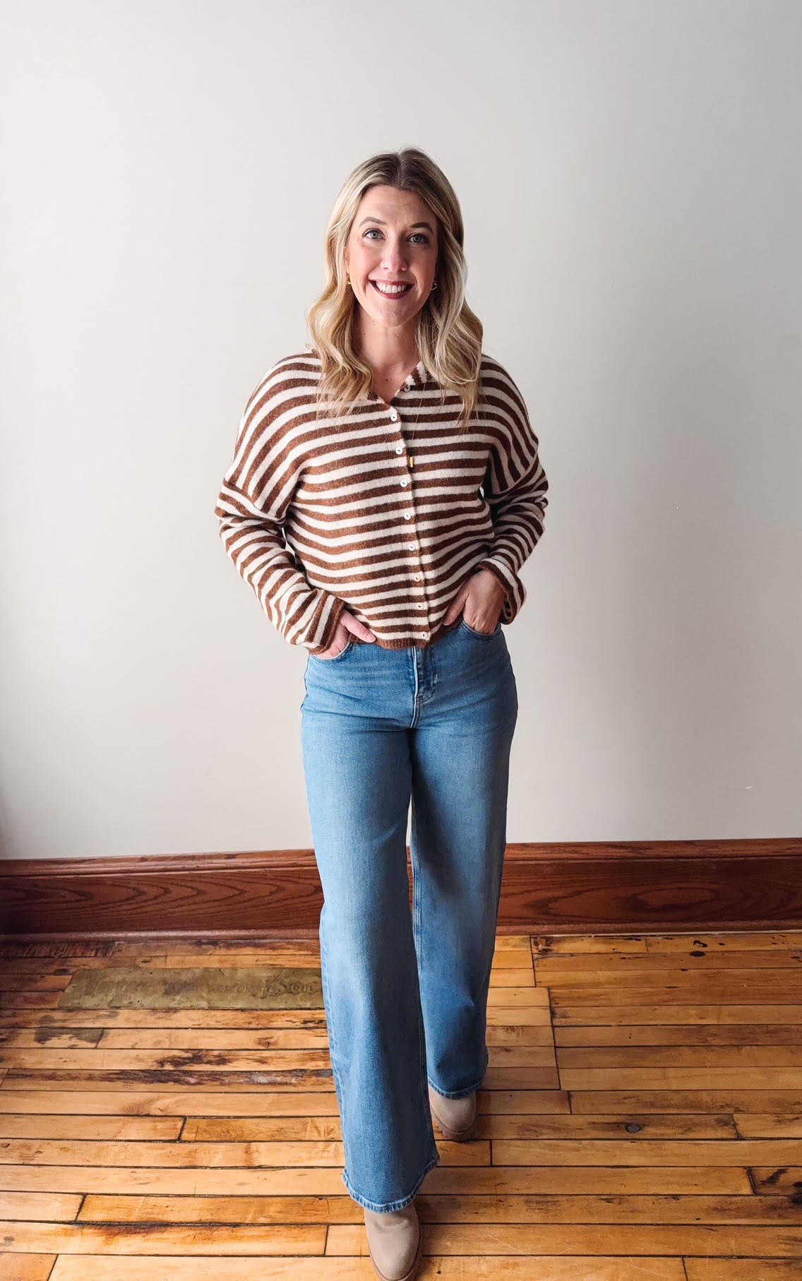 Woman wearing a striped shirt and blue jeans standing on a wooden floor with a white wall background