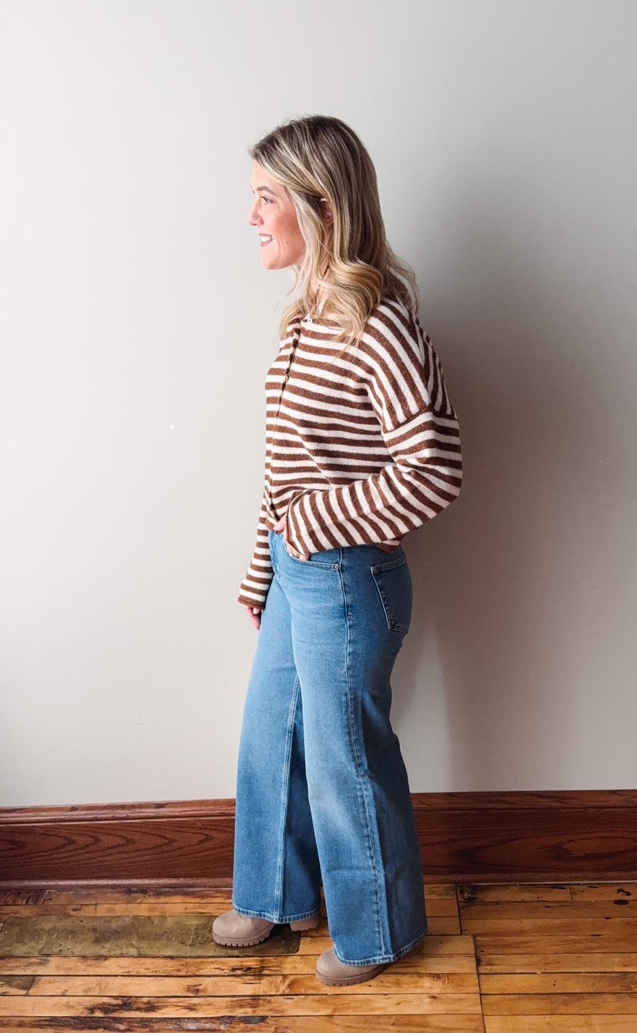 Woman wearing a striped sweater and blue jeans against a plain wall.