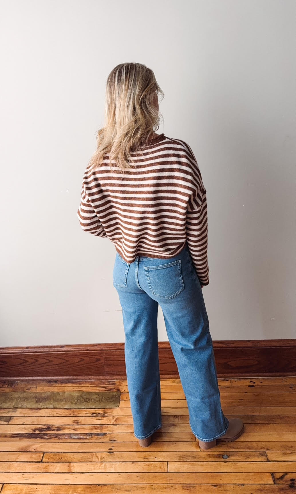 Person wearing a striped sweater and blue jeans standing in a room with wooden flooring and a white wall.