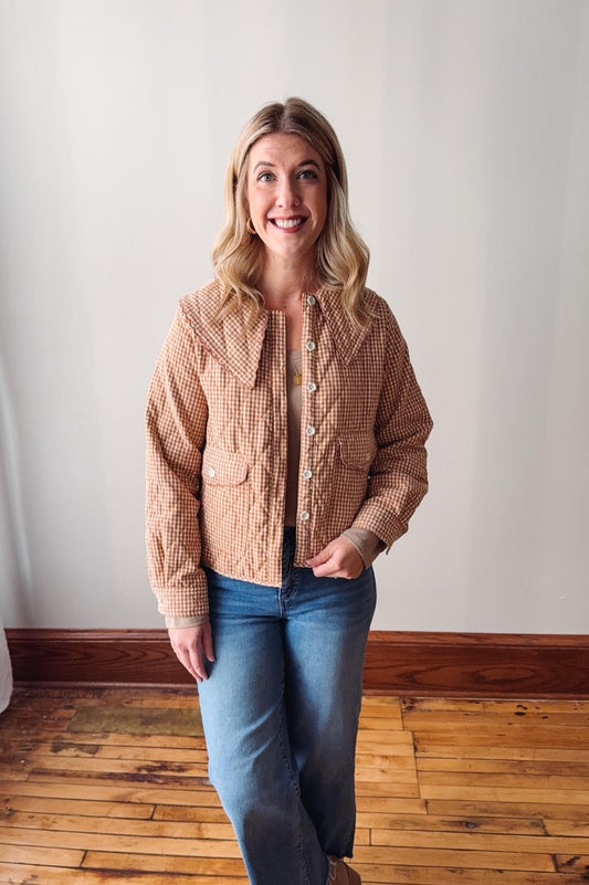 Woman wearing a checkered jacket and jeans standing in a room with wooden flooring and white walls.