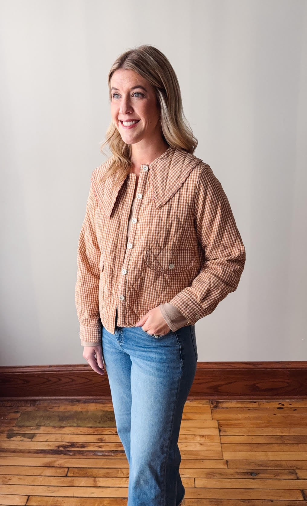 Woman wearing a checkered shirt and jeans standing in a room with wooden flooring and a white wall.