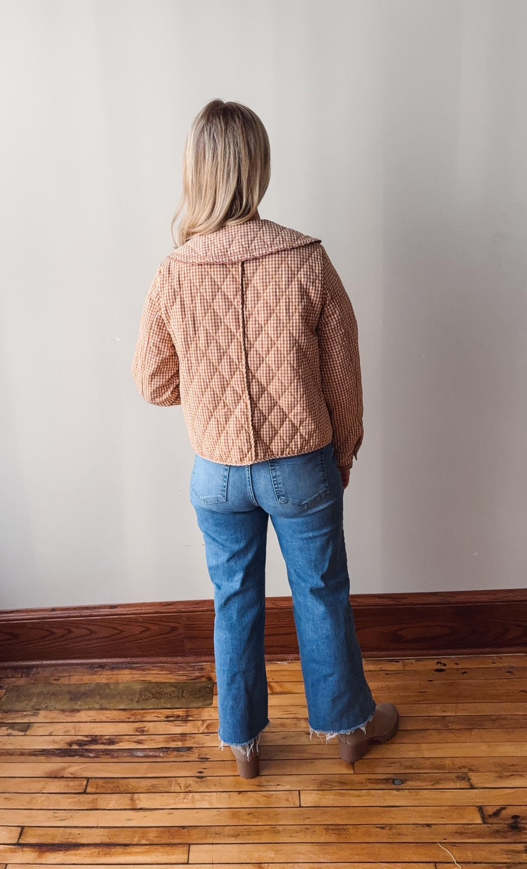 Person wearing a quilted jacket and blue jeans standing on a wooden floor with a plain wall background