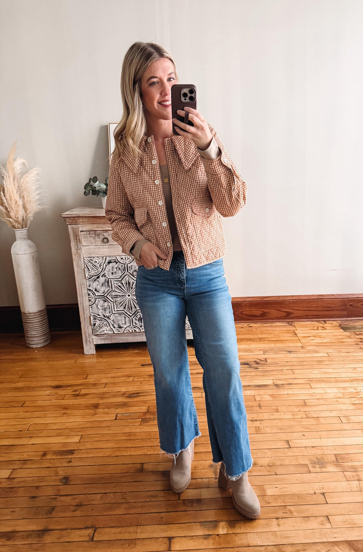 Woman taking a mirror selfie wearing a beige jacket and blue jeans in a room with wooden flooring.