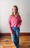 Woman wearing a pink shirt and blue jeans standing in a room with wooden flooring and a white wall.