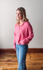 Woman wearing a pink sweater and blue jeans standing against a plain wall.