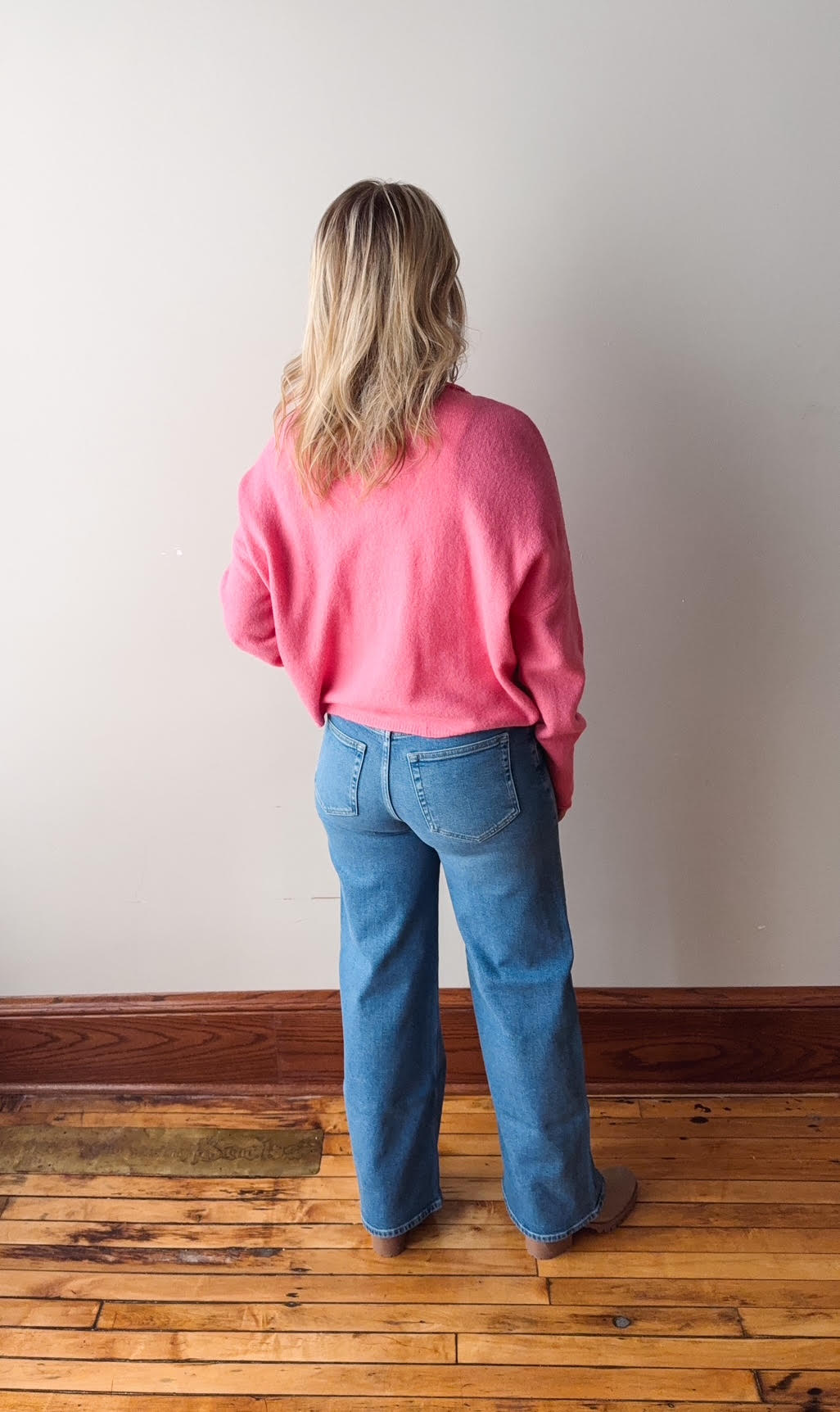 Person wearing a pink sweater and blue jeans standing in a room with wooden flooring and a white wall.