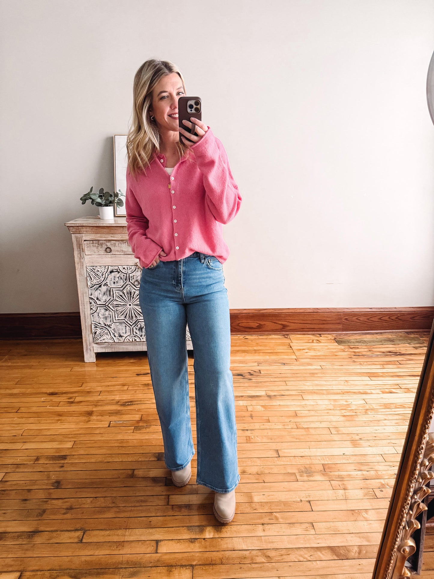 Woman taking a mirror selfie wearing a pink shirt and blue jeans in a room with wooden flooring.
