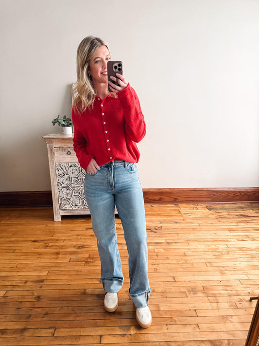Woman taking a mirror selfie wearing a red shirt and blue jeans in a room with wooden flooring.
