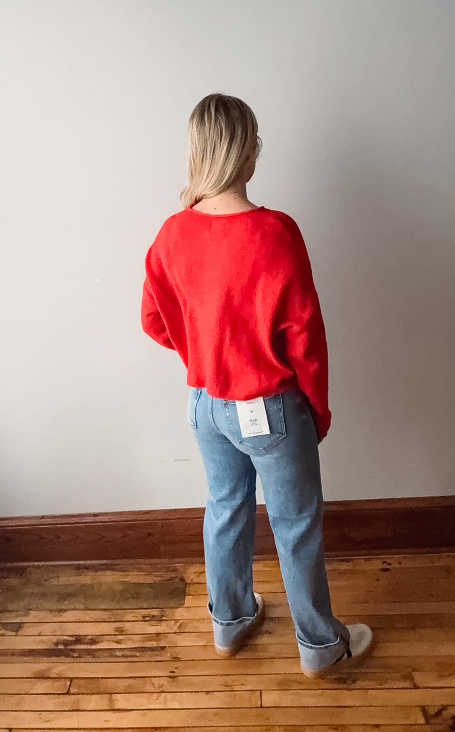 Person wearing a red sweater and blue jeans with a visible tag, standing in a room with wooden flooring and a white wall.
