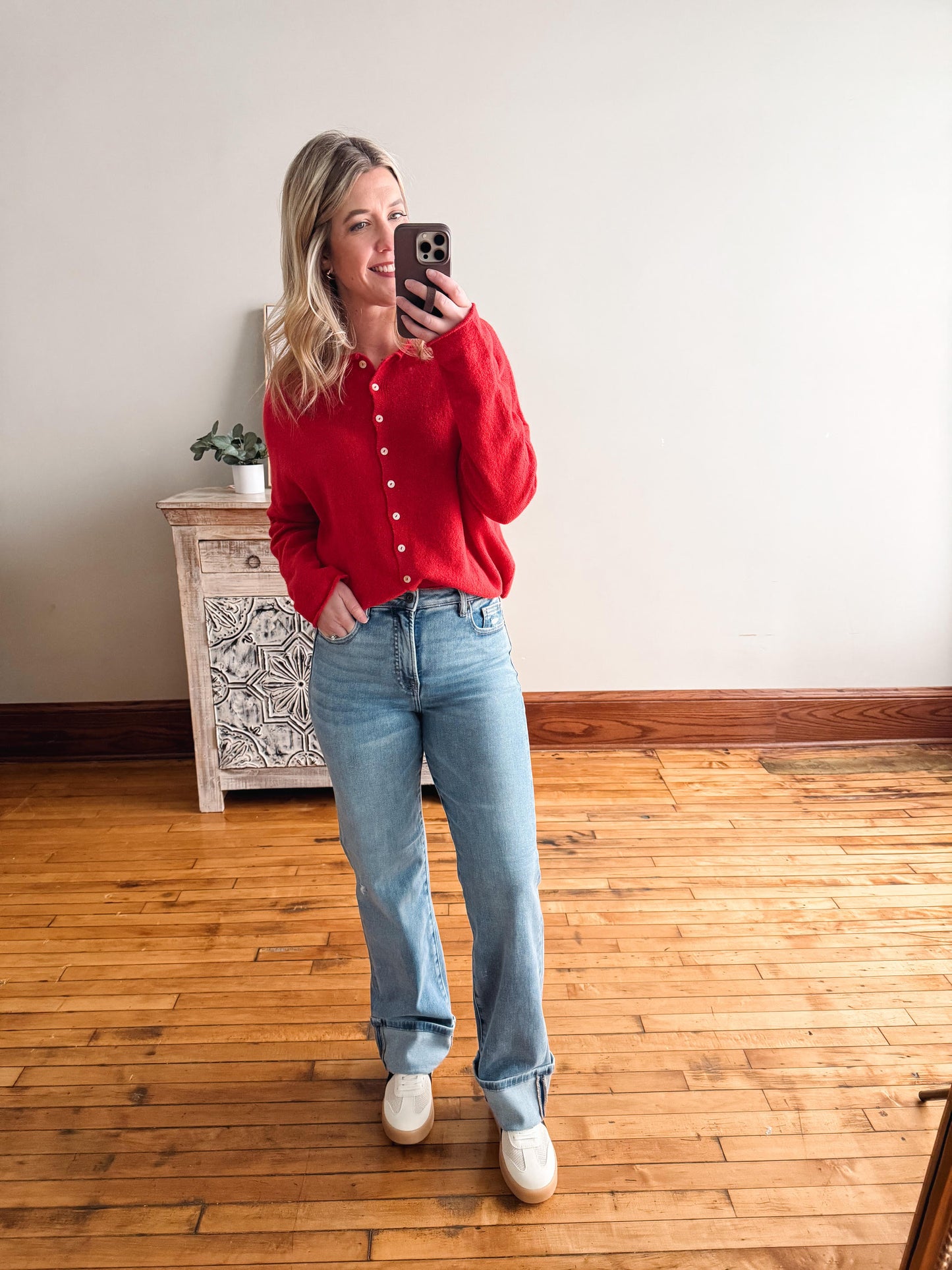 Woman taking a mirror selfie wearing a red shirt and blue jeans in a room with wooden flooring.